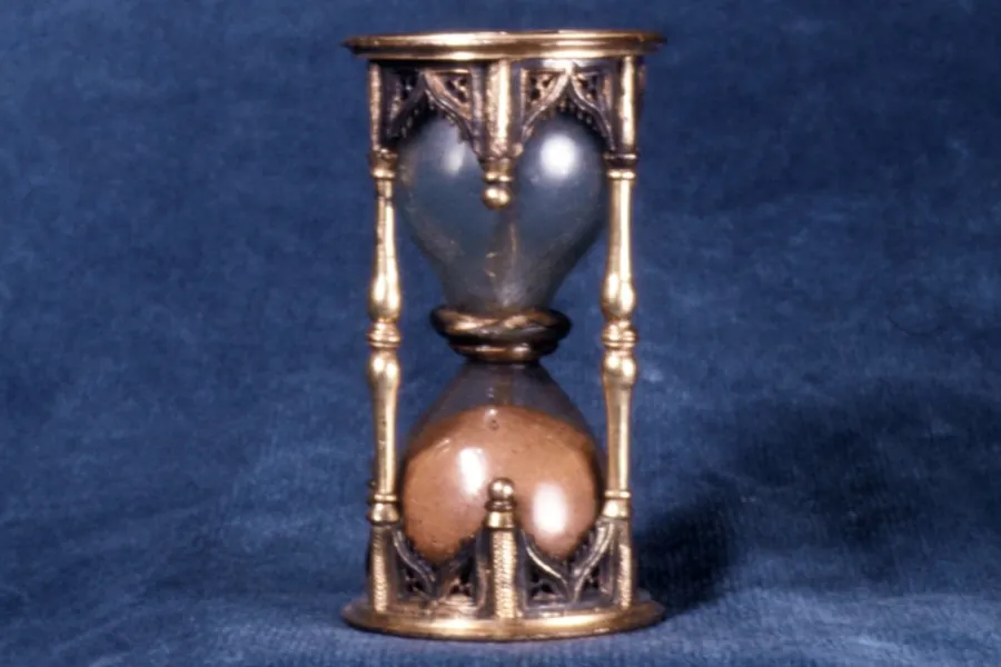 A small brass half-hour sand glass in a gothic-style frame, photographed against blue cloth.